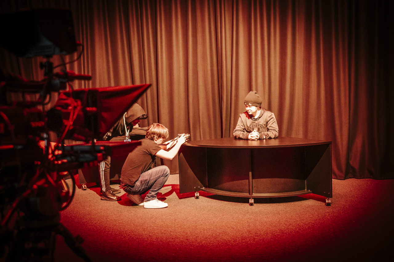 media and film student crouched holding a camera filming a student sat at a news desk