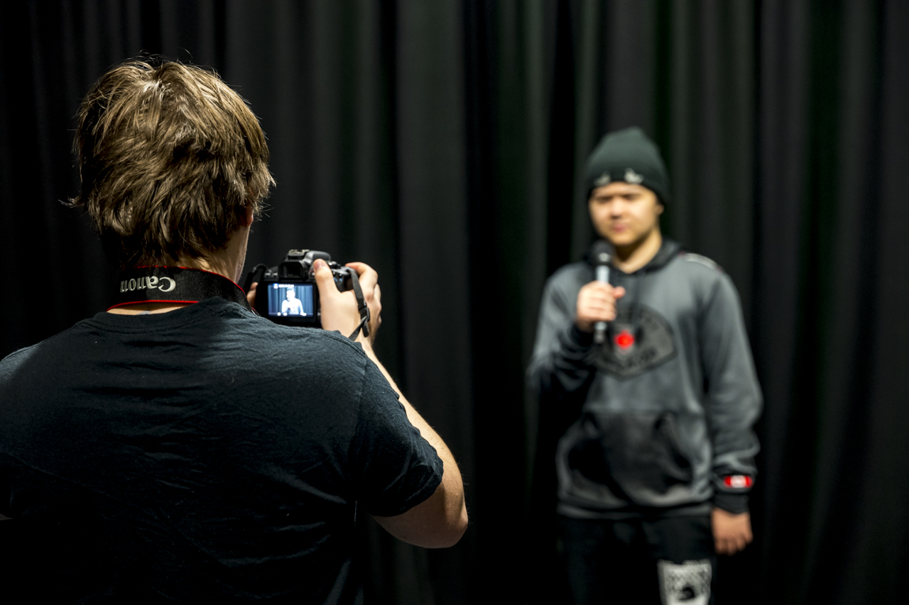 media and film student stood in the foreground with a camera in his hands filming a student holding a microphone