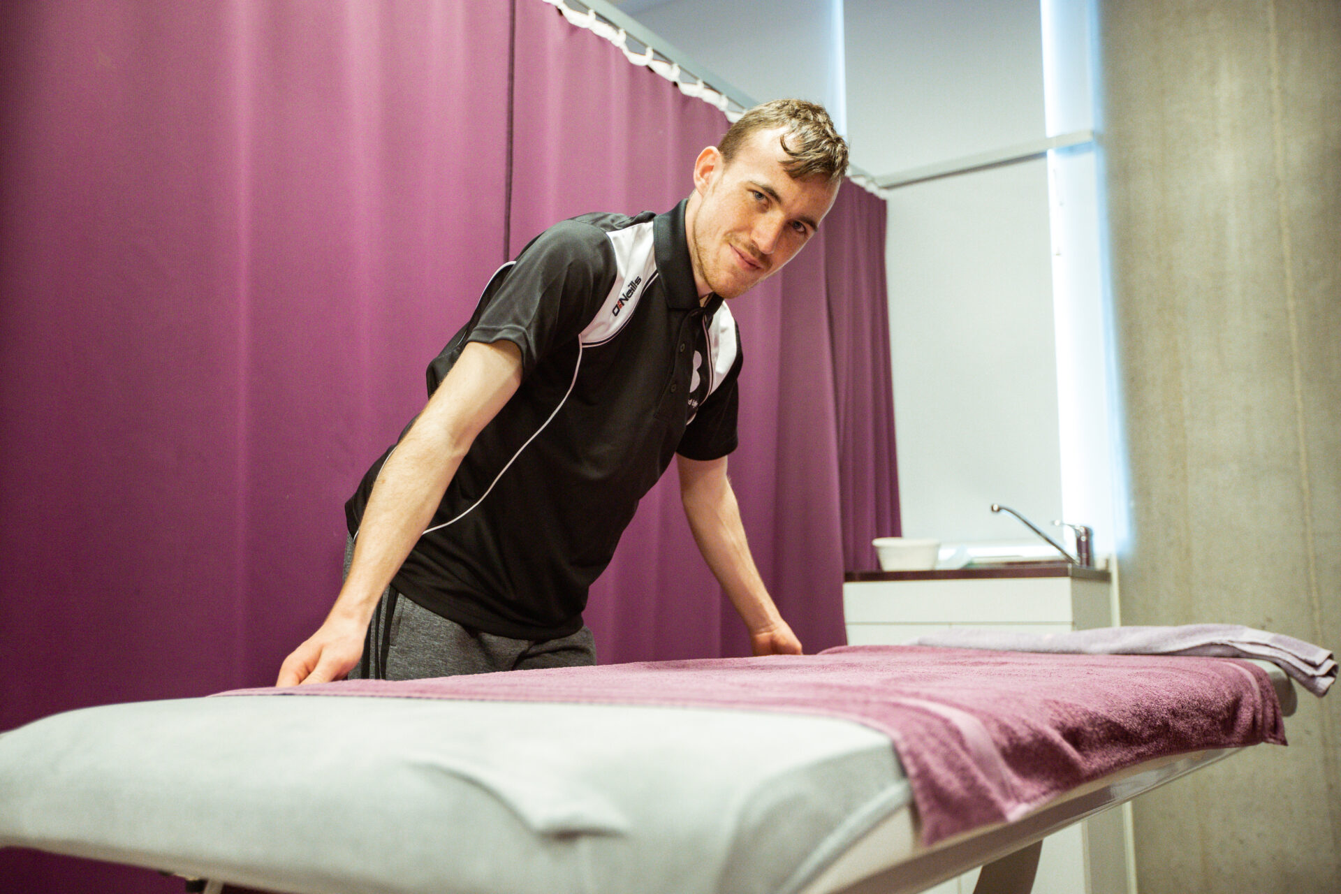 A sports therapist in a black polo shirt is preparing a massage table covered with a purple towel in a treatment room with purple curtains