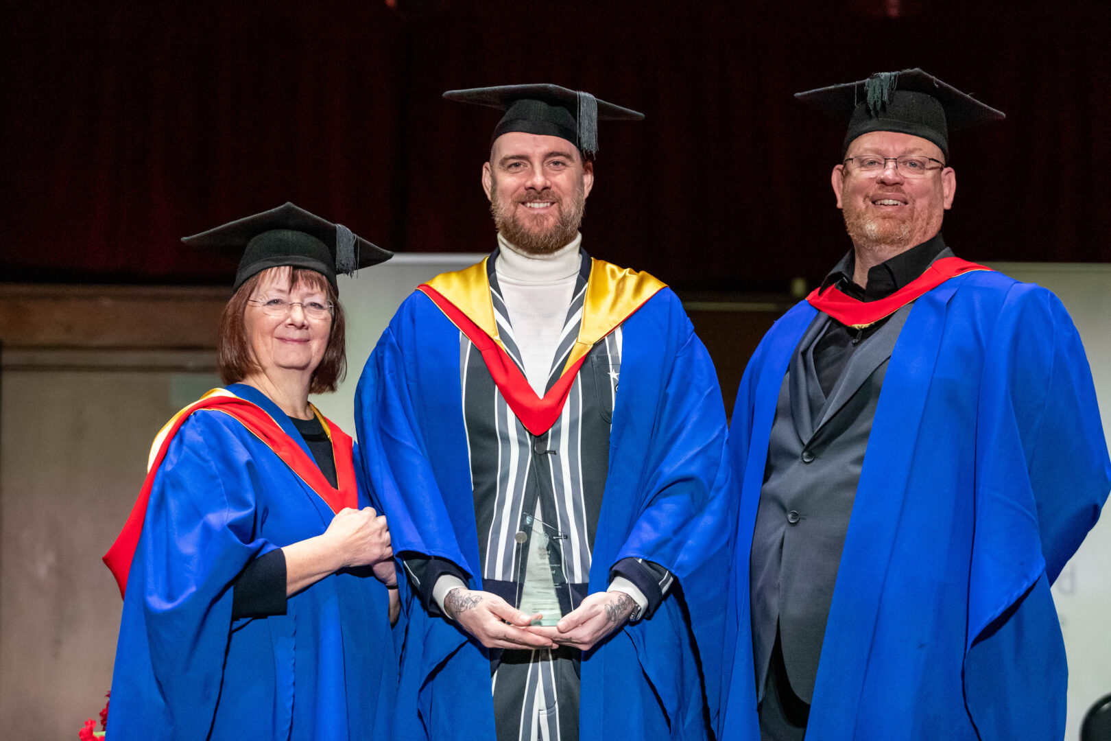 two members of staff with an honorary graduate on stage