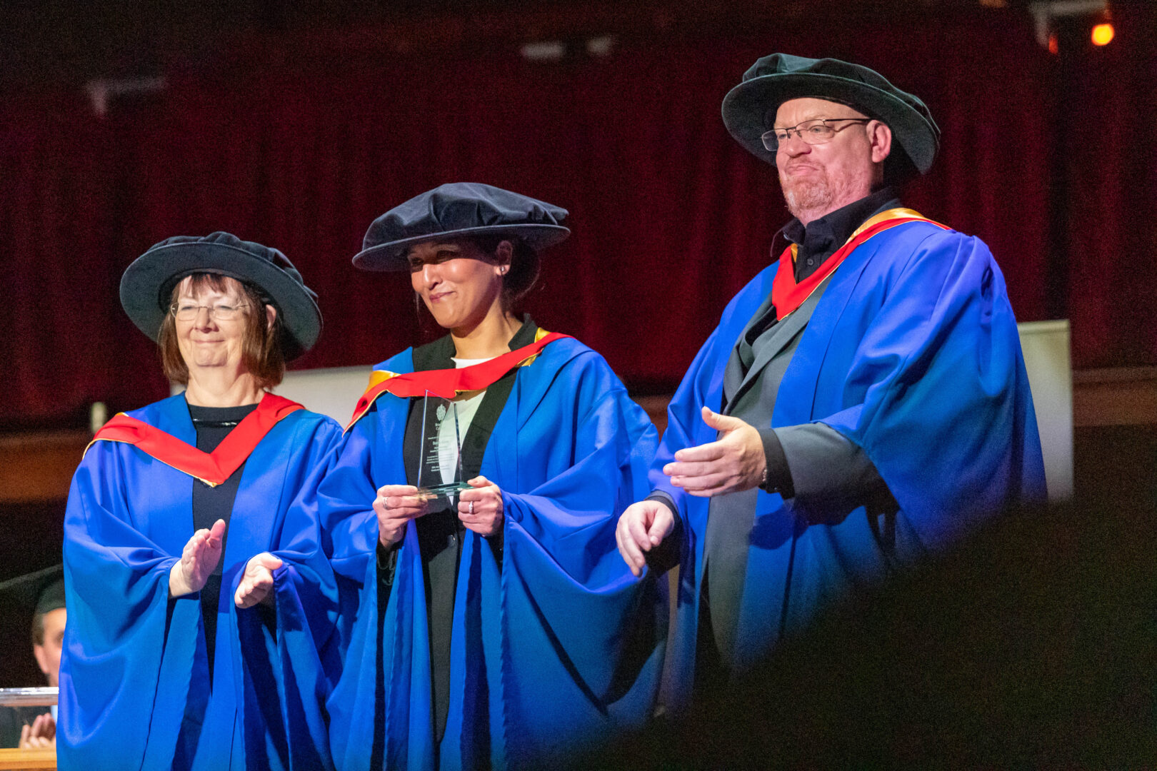 two members of staff with an honorary graduate on stage