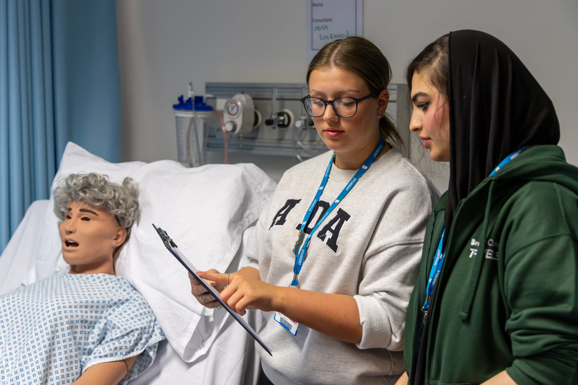 Two female students with clipboard stood beside training doll in our mock clinical ward