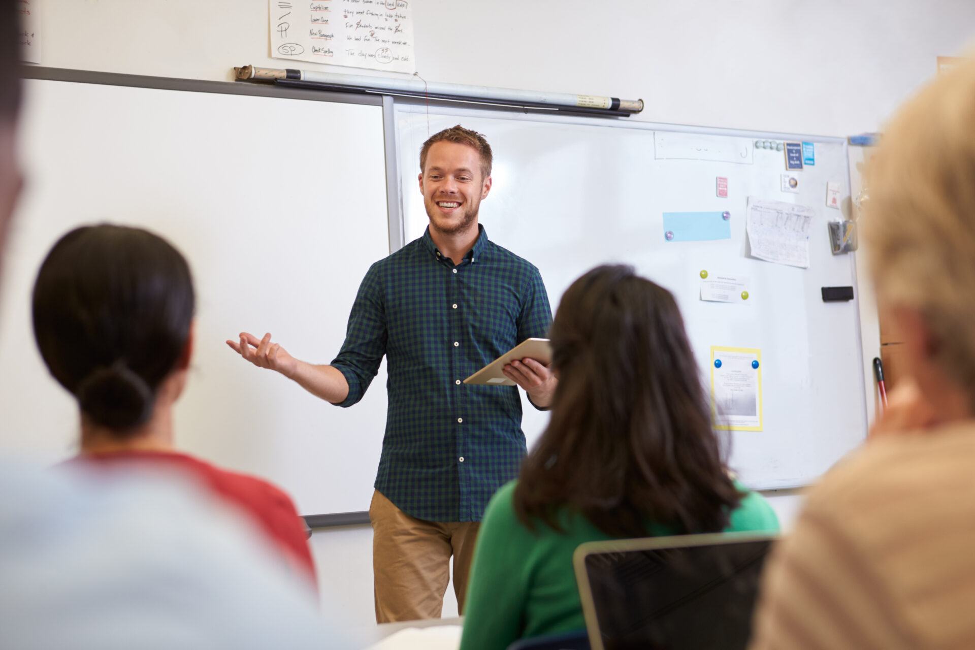 a male teacher stands in front of a class holding a tablet and smiling