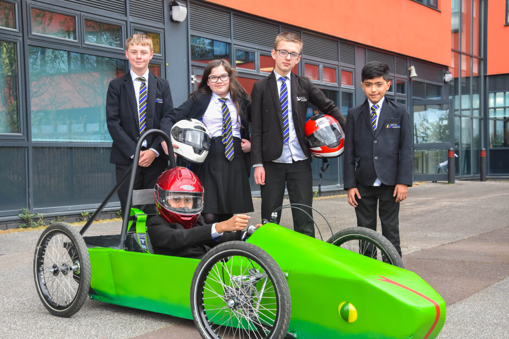 A group of school children stand in front of a school, a child sits in a green electric go kart
