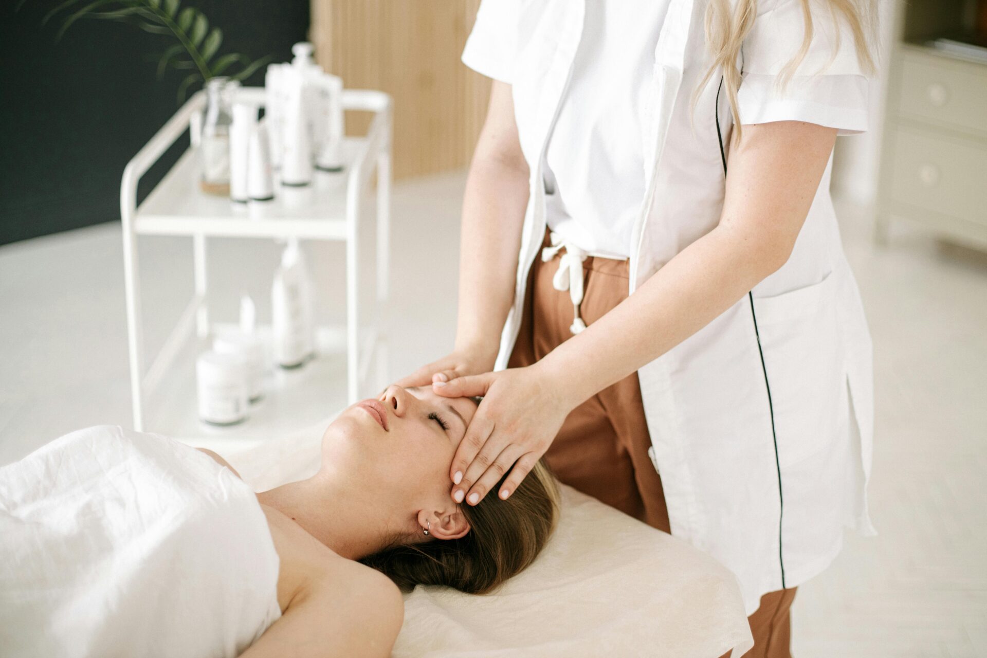 a woman lays on a bed as a beauty therapist wearing a white uniform massages her face