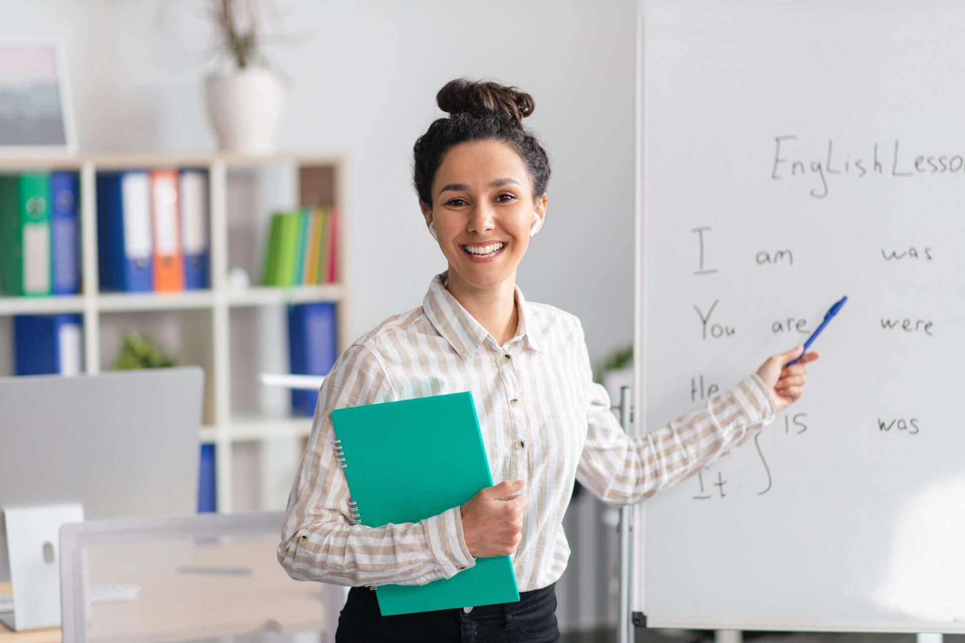 a teacher stands at the front of the class smiling as she points to the whiteboard