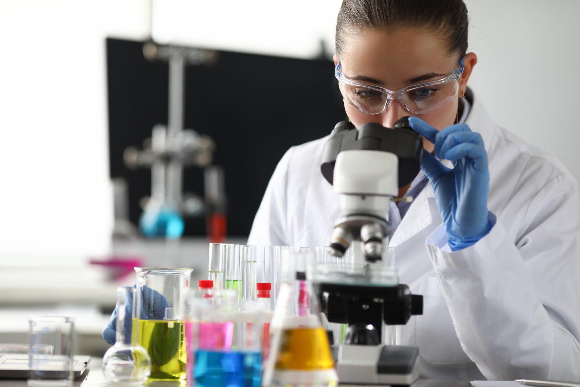 a white female science student wearing safety goggles and gloves looks into a microscope