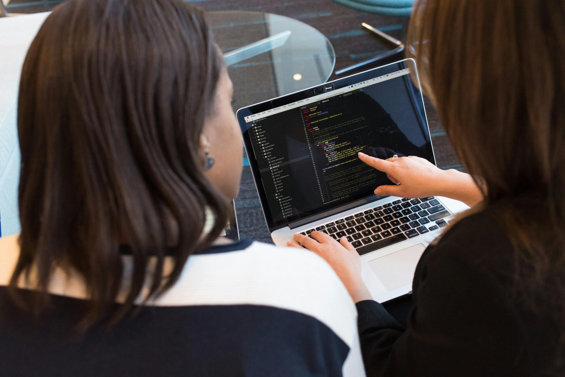 two students sit speaking as they sit and point to a laptop that has coding on the screen