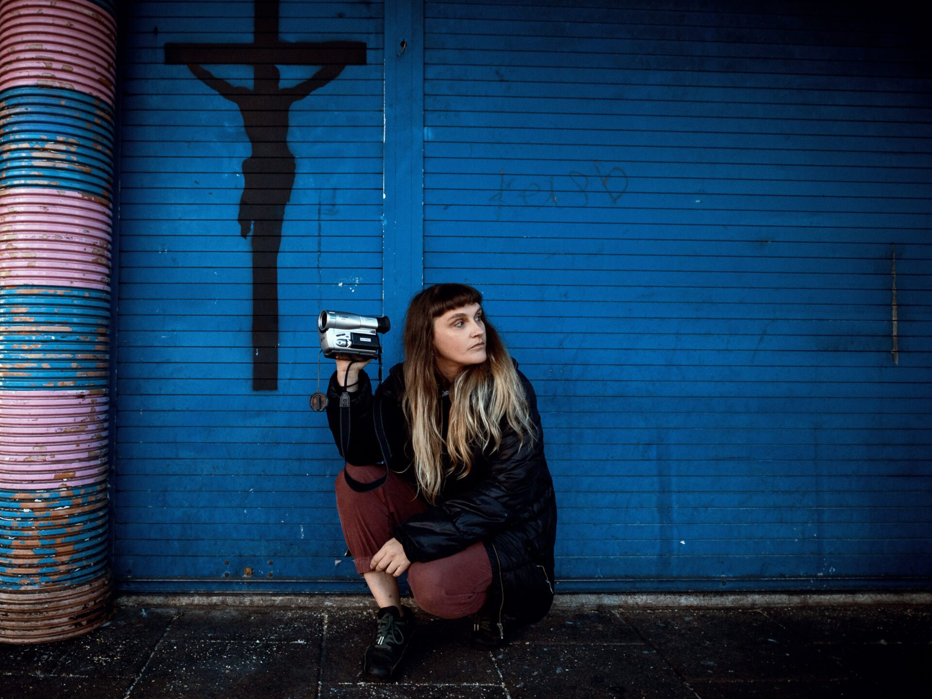 A person crouching against a blue shutter, holding a camcorder, with a silhouette of a cross in the background.