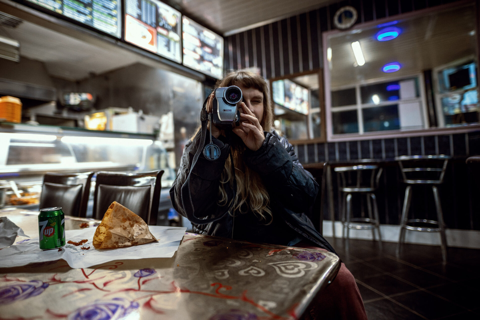 Bradford School of Art Artist-in-Residence, Martha Kean, at a table in a casual eatery, holding a vintage camcorder pointed at the viewer, with food wrappers, and a 7UP can beside them. The background shows a brightly lit counter and menu boards.