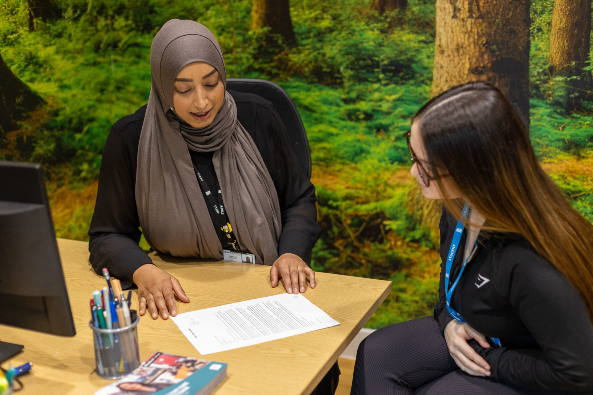 woman in a headscarf sat at a desk talking to female student with long hair wearing glasses