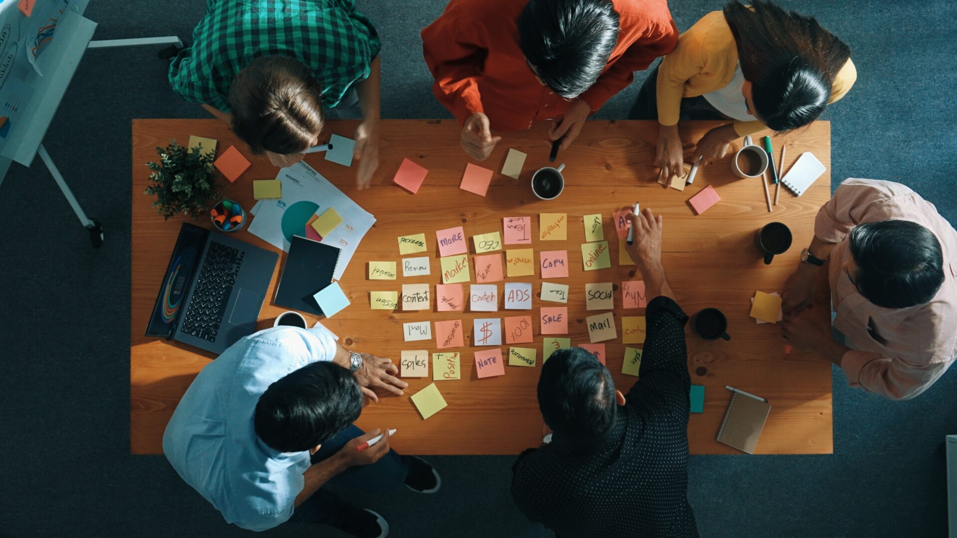 Aerial view of a diverse group of people collaborating around a wooden table covered with colorful sticky notes, a laptop, and coffee cups, brainstorming ideas for a multichannel marketing project.